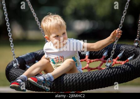 A cheerful little boy swings in the park on a swinging rope swing. Round black and red seat for children's swing. Stock Photo