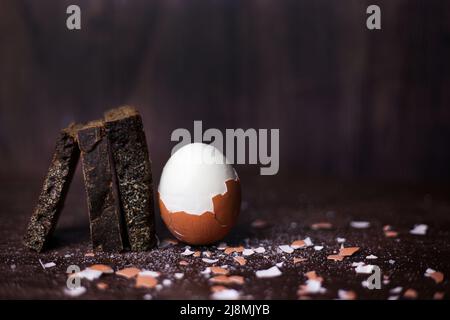 Brown boiled half-peeled egg and pieces of black bread on a dark wooden background with scattered salt and shell Stock Photo