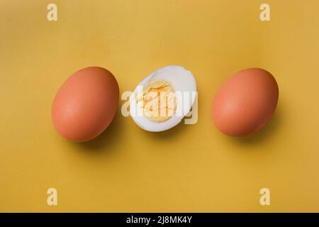 Two whole brown eggs and a boiled egg with yolk cut in half on an isolated light yellow background Stock Photo