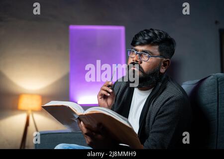 Young Indian beard man thinking while reading book at night - concept of thoughtful and imagination about novel or literature. Stock Photo