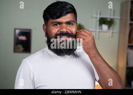 happy young beard man dressing mustache or beard by looking at camera - concept of haircare, relaxation, getting ready and feeling proud for long Stock Photo