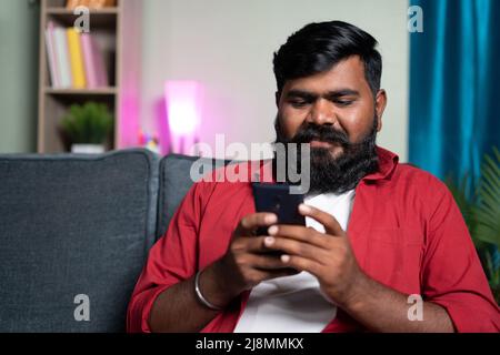 Happy smiling bead man busy using mobile phone on sofa - concpet of social media chatting and surfing internet and reading messages. Stock Photo