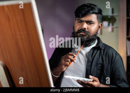 young indian beard artist thinking while painting on canvas board at home - concept of hobbies, leisure activity and professional artist. Stock Photo