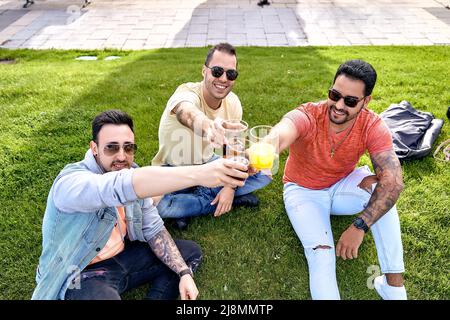 Three friends sitting on the grass, having drinks and toasting while looking at the camera on a sunny day. High quality photo Stock Photo