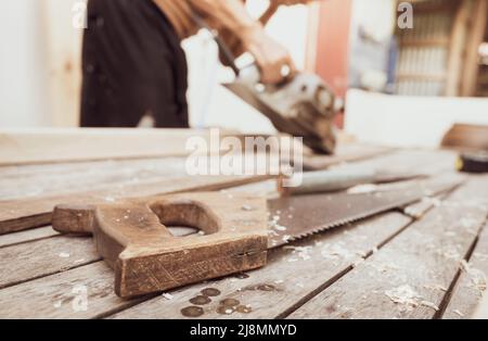Hand saw with a wooden handle on blur carpenter working with electric wood planer. Carpenter tools. Hand saw and sawdust on wood table at outdoor work Stock Photo