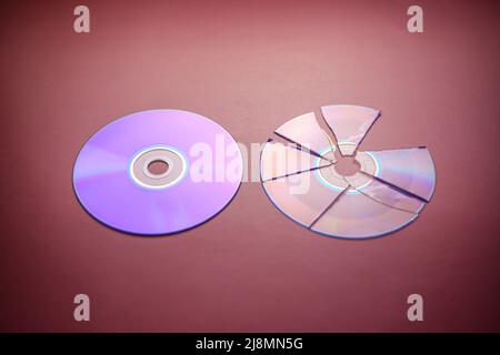Broken compact disc divided into parts and a whole compact disc close-up on a red-burgundy background, complete loss of data Stock Photo