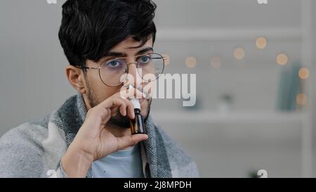 Close-up arabian indian bearded guy with glasses lonely sad sick ill man wrapped in blanket spraying rhinitis spray anti-allergic remedy drug in nose