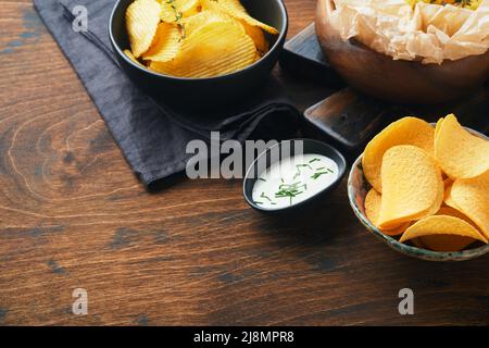 Potato corrugatedchips. Fast food. Crispy potato chips ceramic black bowl with sour cream sauce and onions in wooden stand on old wooden dark backgrou Stock Photo