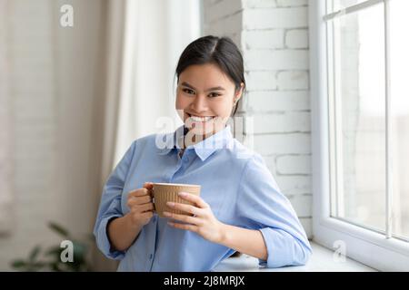 Positive pretty asian woman standing by window, drinking coffee Stock Photo