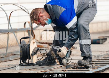 Builder in overalls leans over and cuts off metal sheet with angle grinder and sparks fly. Working man wearing goggles at work on street. Authentic workflow. Stock Photo