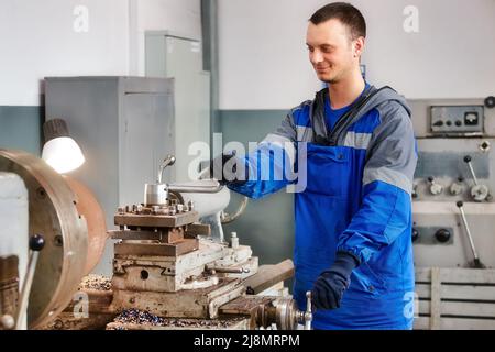 Young turner in overalls works on lathe in workshop. Authentic scene workflow. Caucasian worker recycles metal parts. Stock Photo
