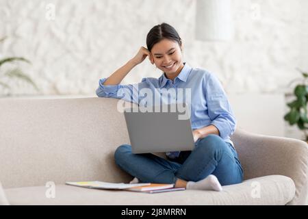 Happy young asian woman sitting on sofa with laptop Stock Photo