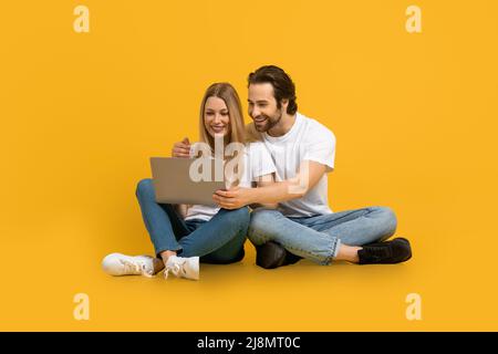 Glad young european man with beard and woman hugs, sit on floor, look at laptop, isolated on yellow background Stock Photo