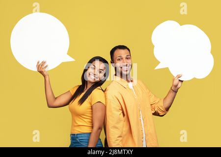 Millennial black couple holding empty speech bubbles on yellow studio background, mockup for design Stock Photo