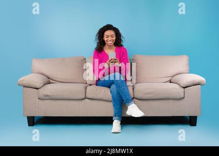 Happy millennial black lady using cellphone, working or studying remotely, sitting on sofa over blue studio background Stock Photo