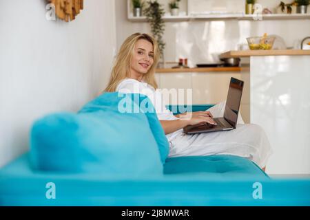 Portrait of blonde beautiful female smiling to camera while working on laptop at home. Stock Photo