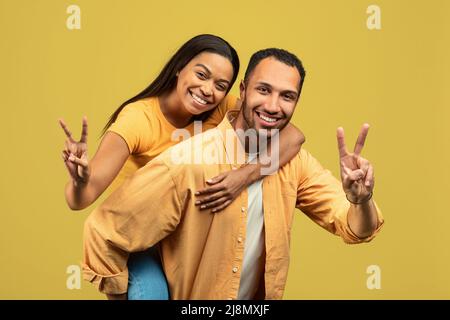 Handsome young African American man giving piggyback ride to his girlfriend, showing peace gesture on yellow background Stock Photo