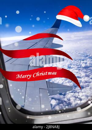 Airplane with red Santa Christmas hat over clouds and blue sky. Christmas happy travel and vacation concept. Stock Photo