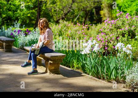 Senior white woman sitting on a stone bench in a park with many flowers. Stock Photo