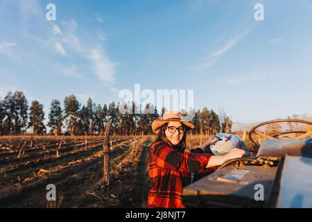 Latin farmer woman getting on an old tractor to drive it, in the middle of her farmland.  Stock Photo