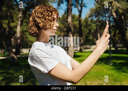 Young redhead woman wearing white tee taking smart phone by phone while standing on city park, outdoor. Stock Photo