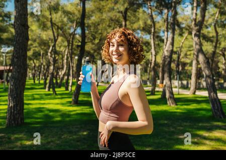 Beautiful happy redhead woman wearing sport bra holding a bottle of energy drink while posing to camera at city park, outdoors. Sportive woman or outd Stock Photo