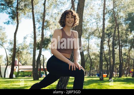 Young redhead happy woman wearing sport bra and black legging warm up or stretching legs before jogging at city park, outdoors. Healthy and sportive l Stock Photo