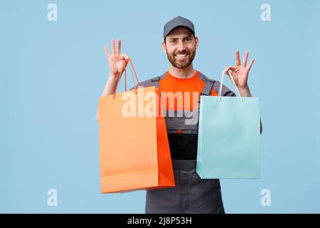 Happy delivery man in uniform holding shopping bags on blue studio background Stock Photo