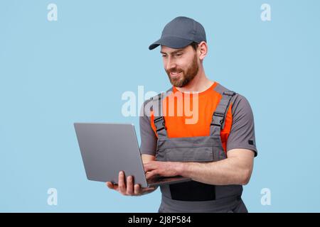 Handsome smiling worker man in uniform work on laptop on blue studio background Stock Photo