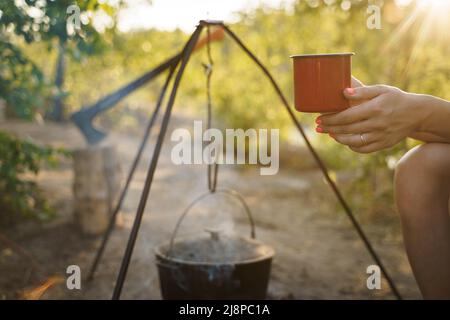 Black large tourist pot hangs over burning flame of campfire in the forest. Outdoor travel. Close up cropped photo. focus on the female hand holding Stock Photo