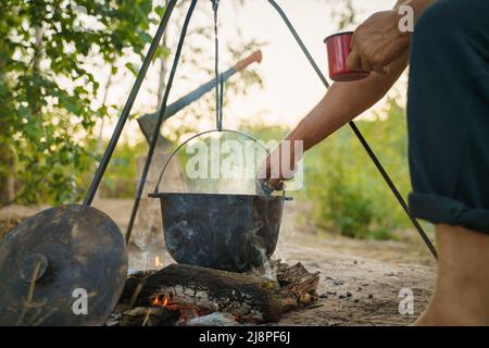 Black large tourist pot hangs over burning flame of campfire in the forest. Outdoor travel. Close up cropped photo. focus on the man's hand holding Stock Photo