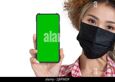 Closed-up female tourist face wareing red plaid shirt with straw hat and face mask holding smartphone shown green sereen on white background Stock Photo