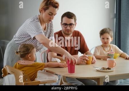Parents with children having meal, breakfast by the table. Mother serving food to family at home. Eating together with kids. Girl in high chair eating Stock Photo