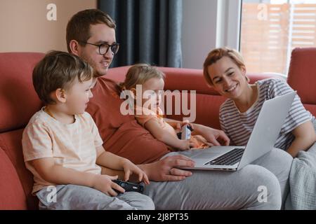 Big happy family watching movies playing games on laptop. Mother father kids spending time with gadgets on sofa Children with parents looking on Stock Photo