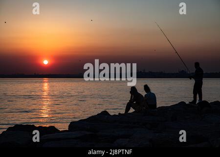 A silhouette of a man fishing and people sitting on the cliffs is seen as  the sun goes down. A man fishing at sunset and people watching the scenery  on the rocks