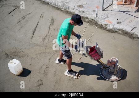 Aerial view of man builder using troweling machine while screeding floor. Worker finishing concrete surface with floor screed grinder machine at construction site on the roof of residential building. Stock Photo