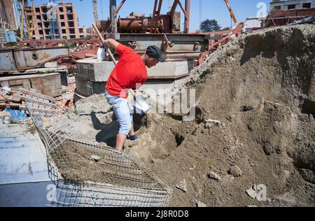 Side view of floor screeding contractor shoveling sand-cement mix outdoors at construction site. Male worker with shovel in his hands preparing cementitious material for floor screed. Stock Photo