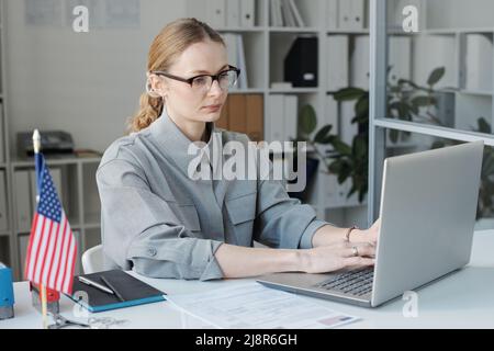 Horizontal medium shot of female visa service specialist sitting at desk working with application documents on laptop Stock Photo