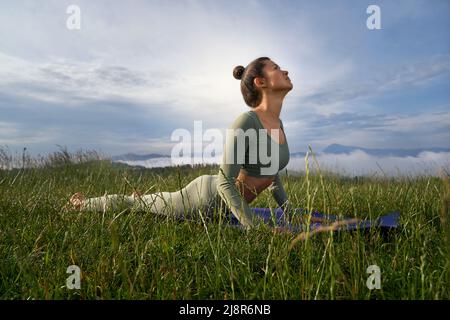 Brunette woman doing Surya Namaskar in mountains among green grass, looking at sky. Pretty female have practice in morning, standing in Bhujangasana, Stock Photo