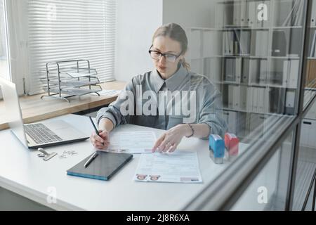 Through glass wall shot of mature Caucasian woman sitting at desk in modern office working with visa application papers Stock Photo