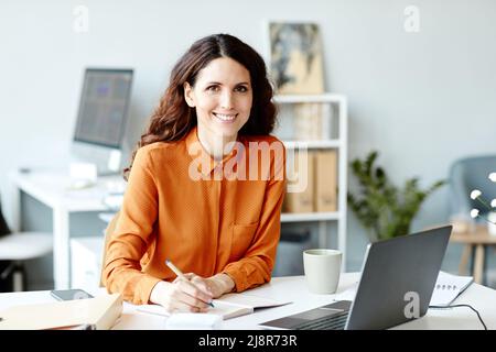 Horizontal medium portrait of joyful young adult woman working in office making notes in notebook smiling at camera Stock Photo