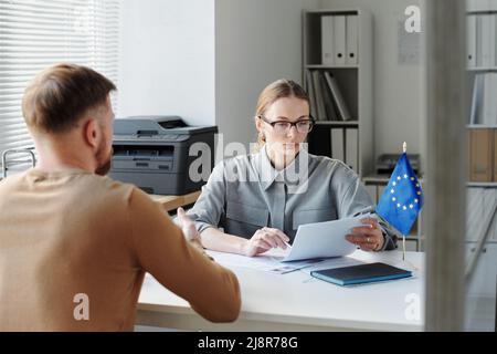 Visa service specialist sitting at table in front of man looking through documents checking data during appointment Stock Photo