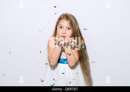 Happy Birthday. Beautiful little girl inflates confetti on a white background. The child is wearing a white dress and a blue ribbon. Big blue eyes and long blond hair. Happy New Year. Copy Space Stock Photo