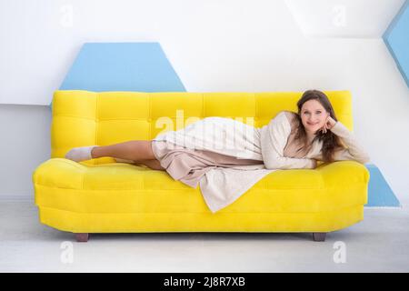 Young smiling caucasian woman 20 y.o. lies on a yellow couch, look at camera, in bright interior with copy space. Stock Photo