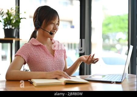 Cheerful Asian millennial female online tutor having an online class, tutoring her students on online platform, remote working in the cafe. Stock Photo