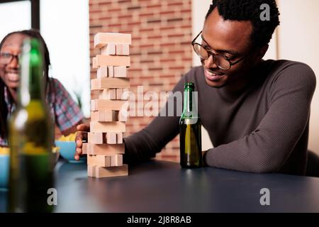 Happy man with beverage on table carefully removing wooden block from wood tower. Smiling person sitting at home in living room with close friends while playing society games together. Stock Photo