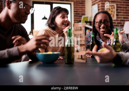 Diverse group of friends laughing together while discussing and playing society games at home. Multiethnic people sitting at table in living room while enjoying fun leisure activity with wood blocks. Stock Photo