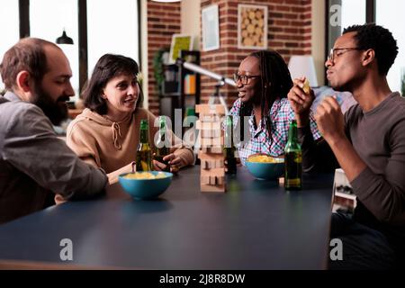 Multiethnic friends sitting at home in living room while talking and playing with wood blocks. Multiracial group of people sitting at table while discussing and enjoying leisure activity. Stock Photo
