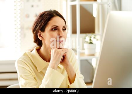 Pensive young adult woman wearing cream beige shirt working in office watching presentation or video on computer Stock Photo