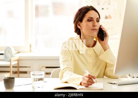 Horizontal medium portrait of Caucasian female CEO sitting at table looking at computer monitor talking a phone with colleague and making notes Stock Photo